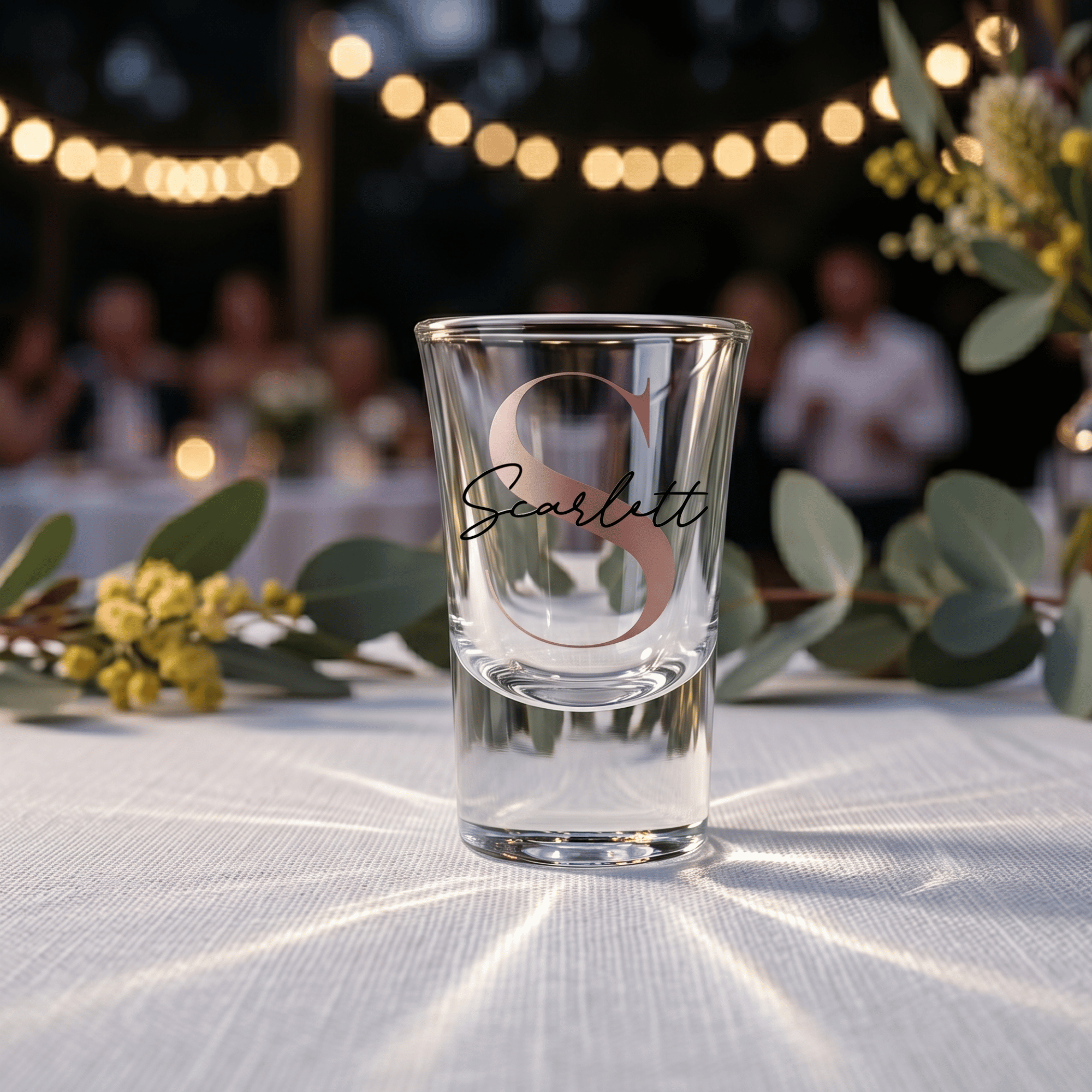 Clear shot glass with 'Scarlett' engraving on a table with decorative elements and blurred people in the background.