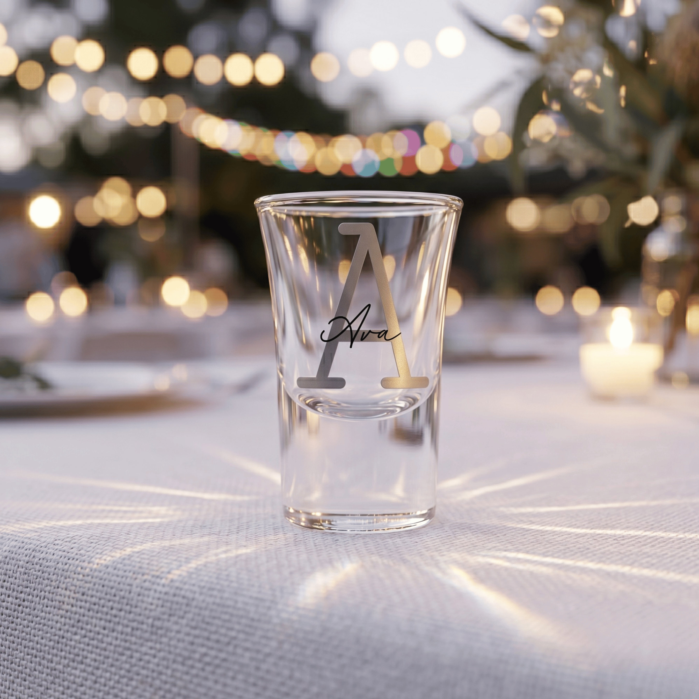 Clear shot glass with engraved letter 'A' on a table with blurred lights in the background