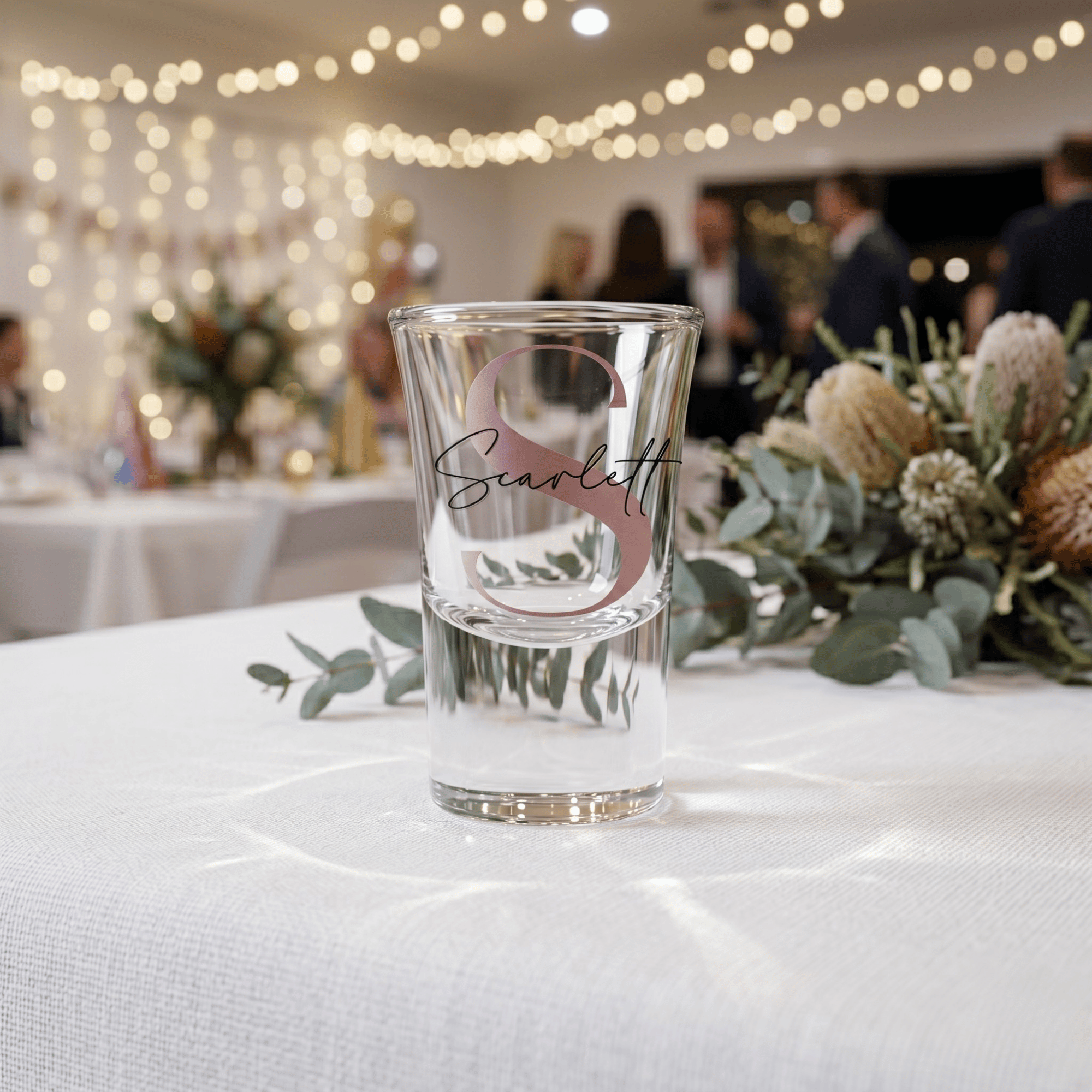 Clear shot glass with 'Scarlett' engraving on a table with decorative elements and blurred background