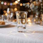 Clear shot glass with '18 Isabella' text on a table with blurred lights and flowers in the background