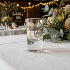 Clear glass with gold number '18' and name on a table with floral decorations