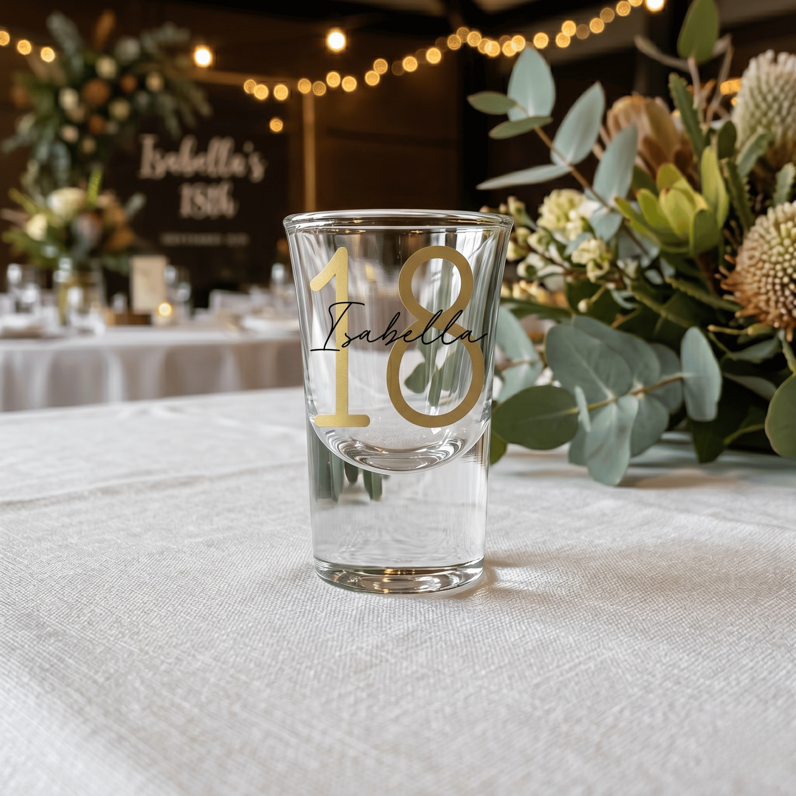 Clear glass with gold number '18' and name on a table with floral decorations