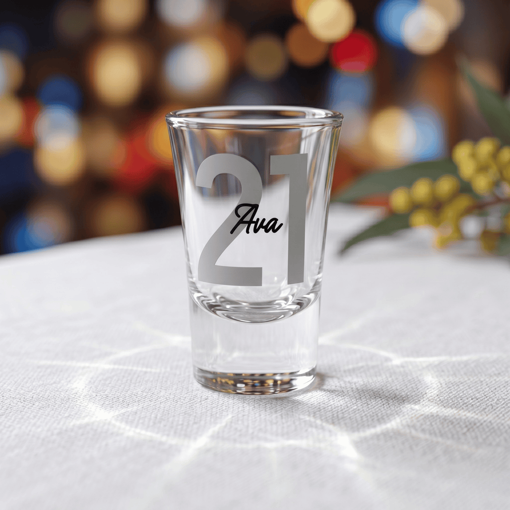 Clear shot glass with '21 Ava' engraving on a white surface with blurred colorful background