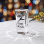 Clear shot glass with '21 Ava' engraving on a white surface with blurred colorful background