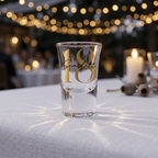 Clear shot glass with gold text on a tablecloth with blurred lights in the background
