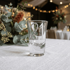 Clear shot glass with engraved text on a table with decorative flowers and blurred background