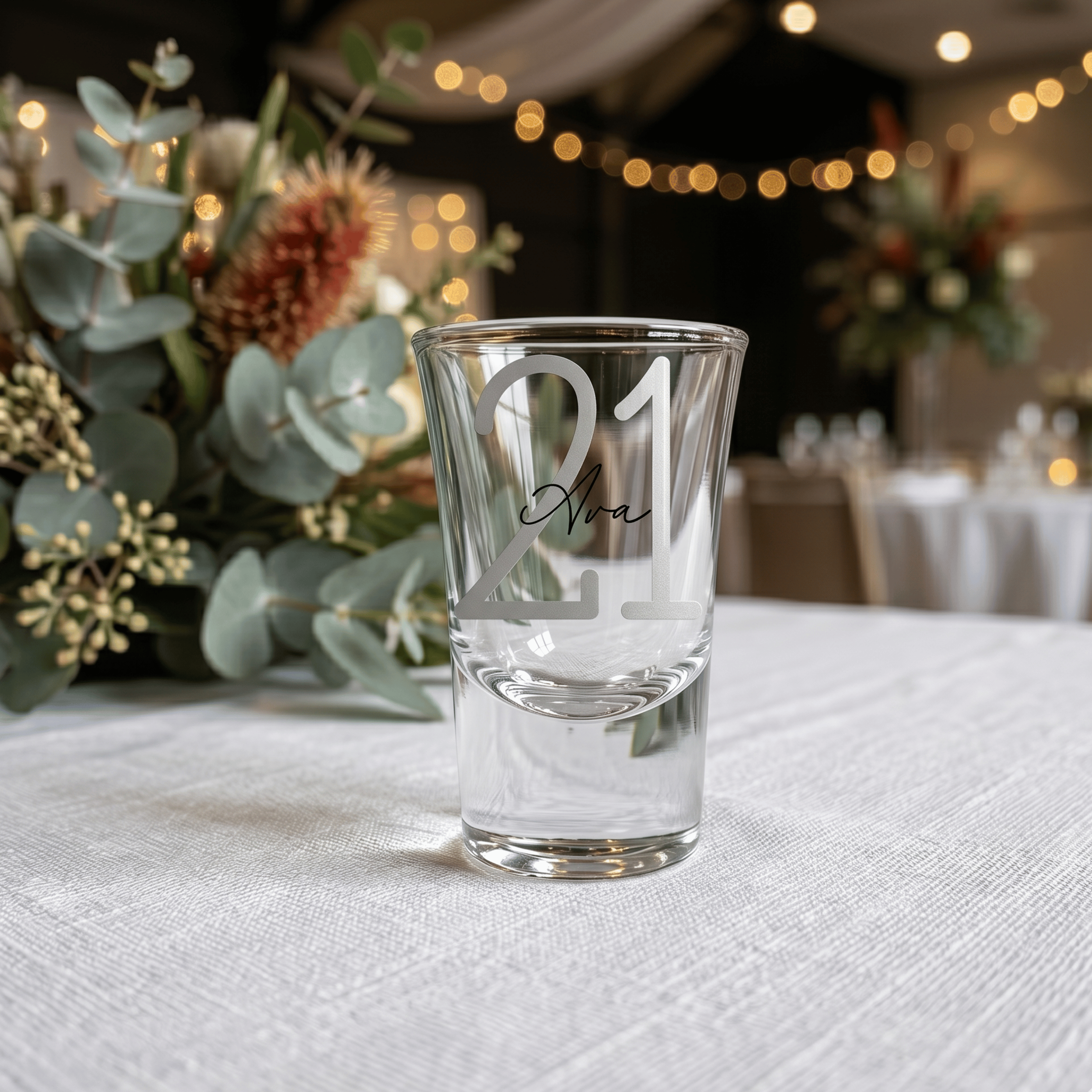 Clear shot glass with engraved text on a table with decorative flowers and blurred background