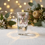 Clear shot glass with 'Scarlett' and '30' on a table with decorative lights and flowers.