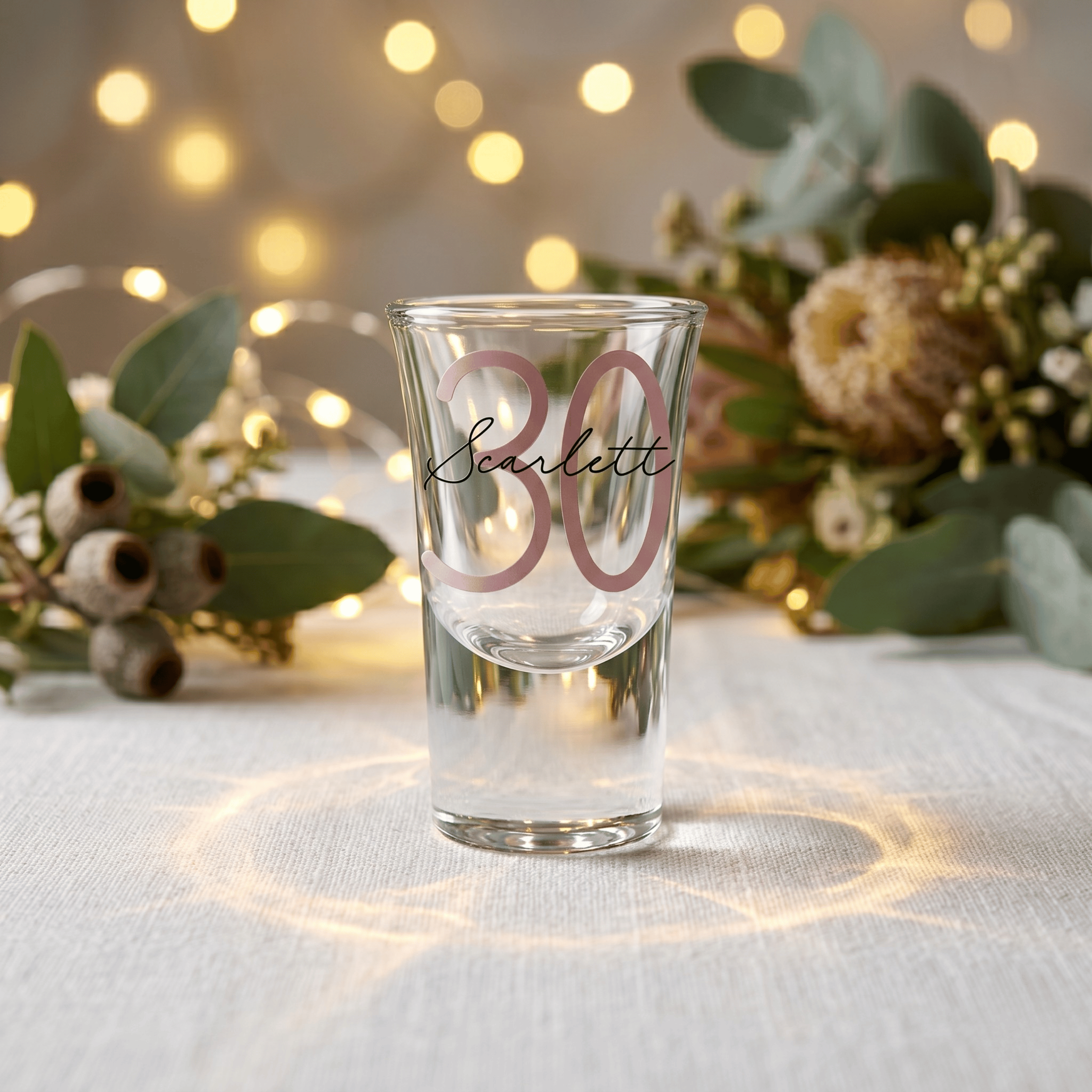 Clear shot glass with 'Scarlett' and '30' on a table with decorative lights and flowers.