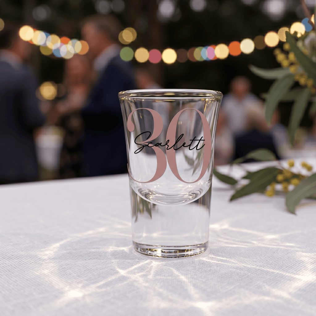 Clear glass with '30 Scarlett' text on a tablecloth with blurred people and lights in the background
