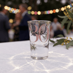 Clear glass with '30 Scarlett' text on a tablecloth with blurred people and lights in the background
