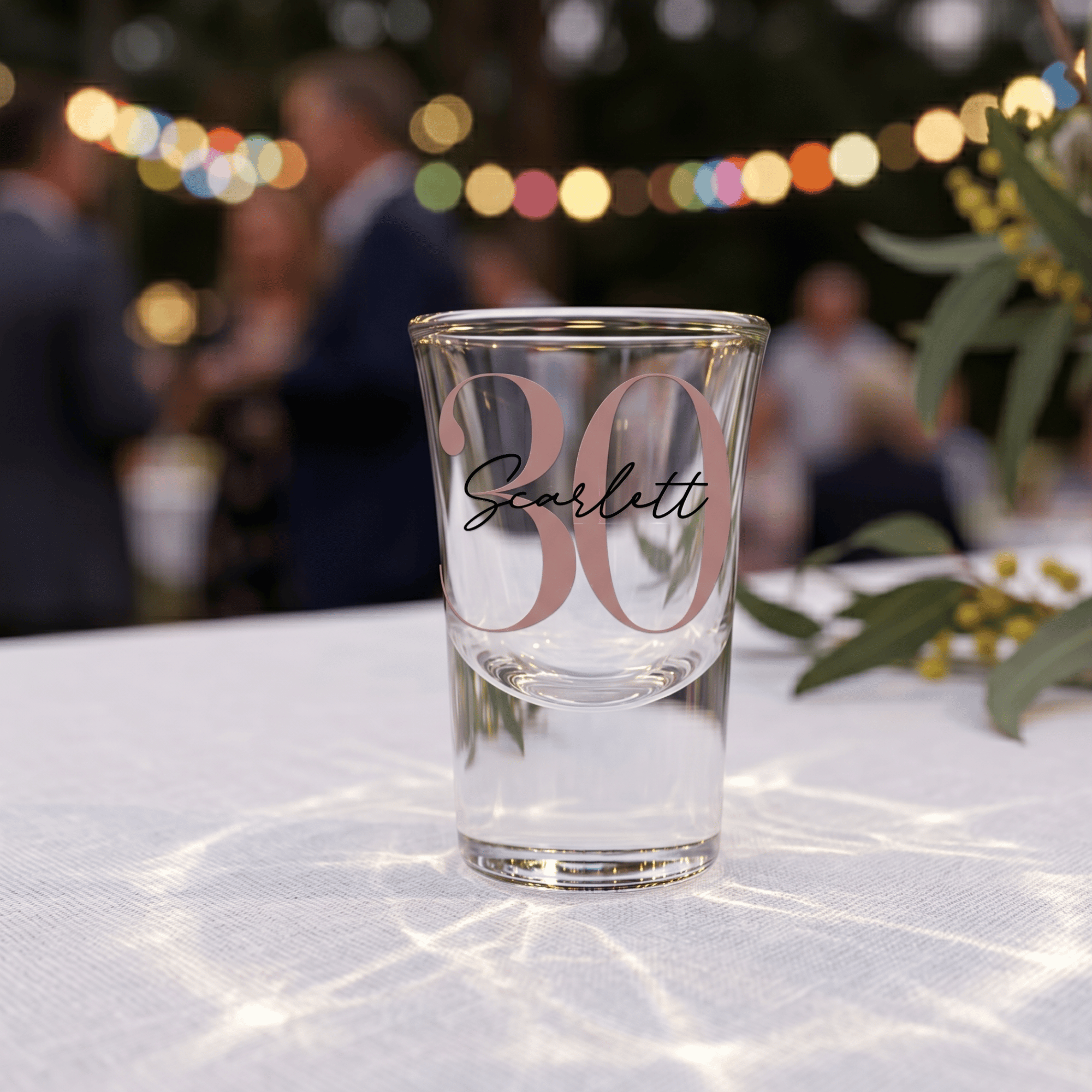 Clear glass with '30 Scarlett' text on a tablecloth with blurred people and lights in the background