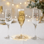 Three champagne flutes with personalized numbers on a table with blurred lights and flowers in the background.