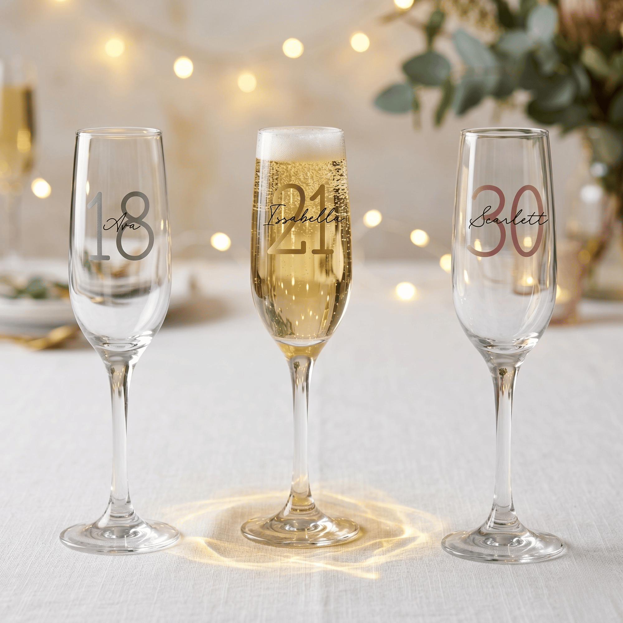 Three champagne flutes with personalized numbers on a table with blurred lights and flowers in the background.