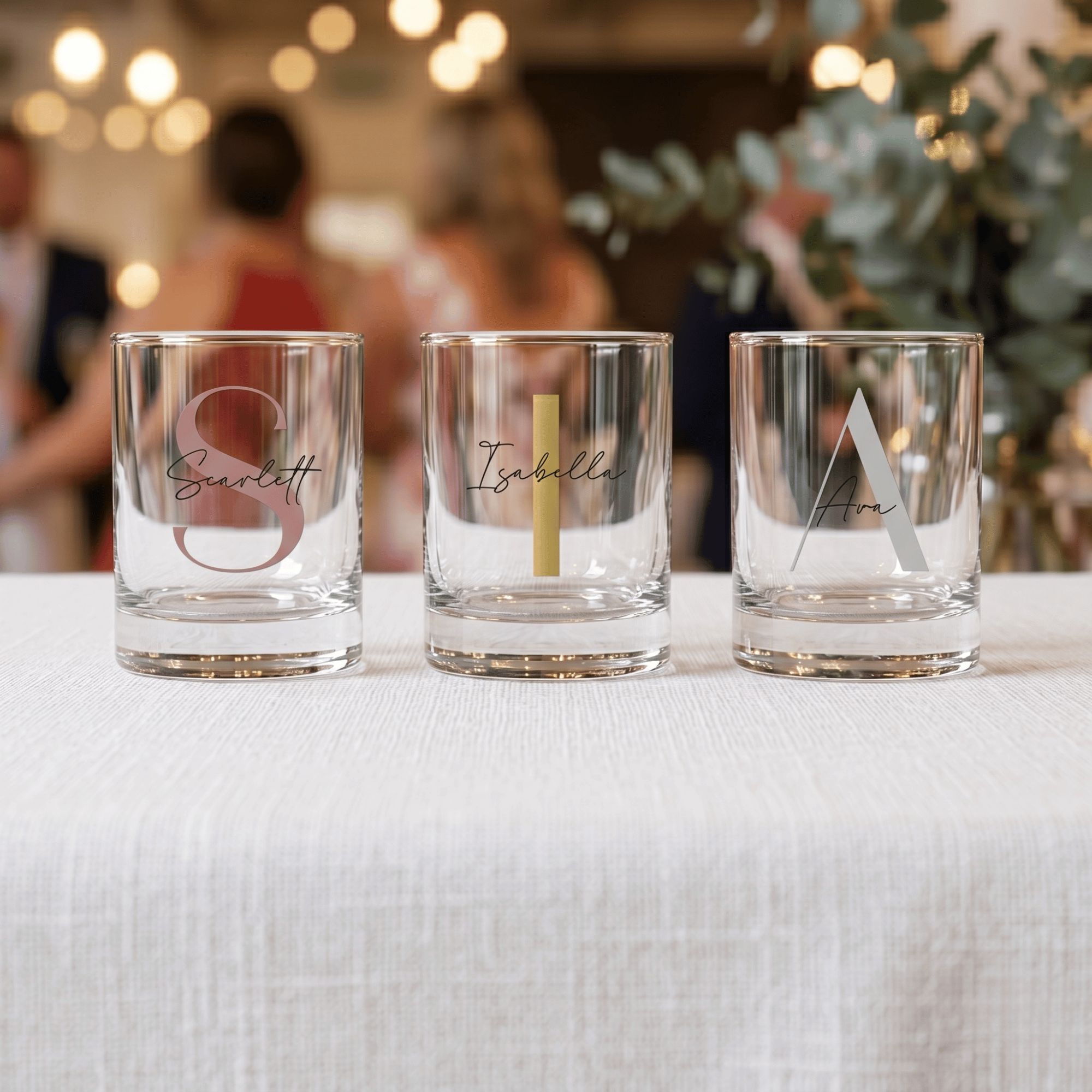 Three personalized glass tumblers on a table with blurred people and decor in the background