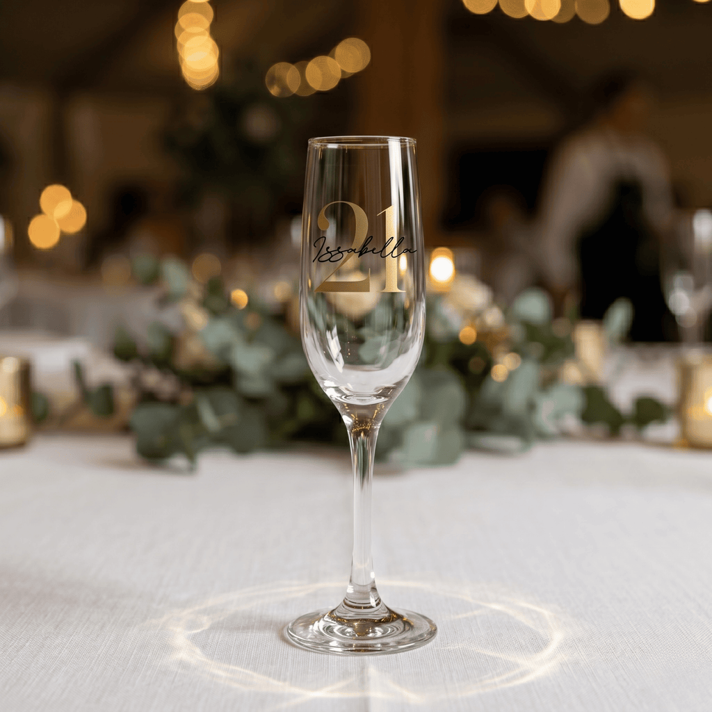 Champagne flute with engraved number 21 on a table with decorative lights and greenery