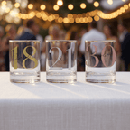 Three clear glasses with numbers and names on a tablecloth with blurred people and lights in the background