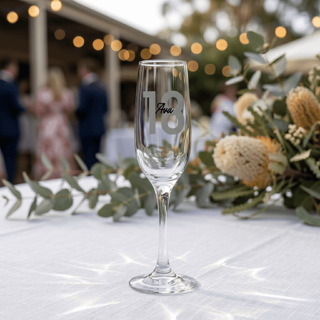 Champagne flute with 'Ava 18' engraving on a table with floral decorations and blurred people in the background
