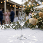 Champagne flute with 'Ava 18' engraving on a table with floral decorations and blurred people in the background