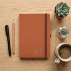 Brown notebook with 'K.S' initials on a wooden surface with stationery items.