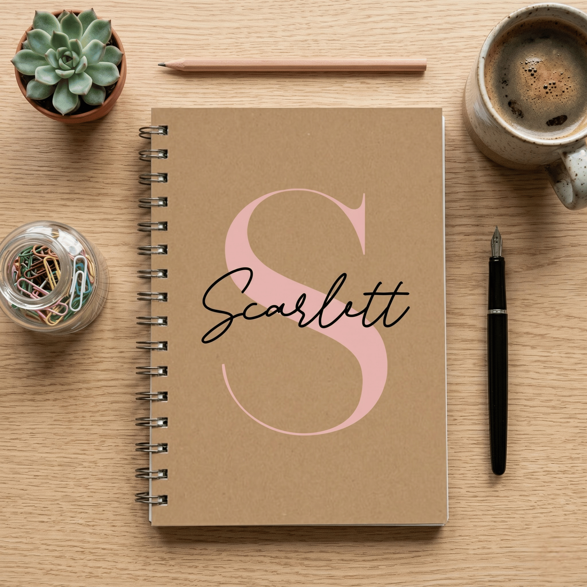 Notebook with 'Scarlett' on a wooden surface with a cup of coffee, pen, and plant.
