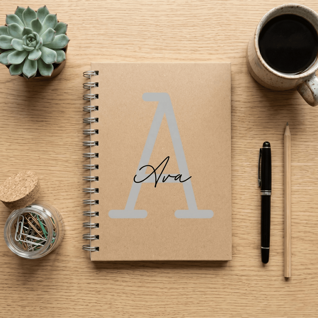 Notebook with 'Ava' on a wooden surface with a plant, mug, and pen.