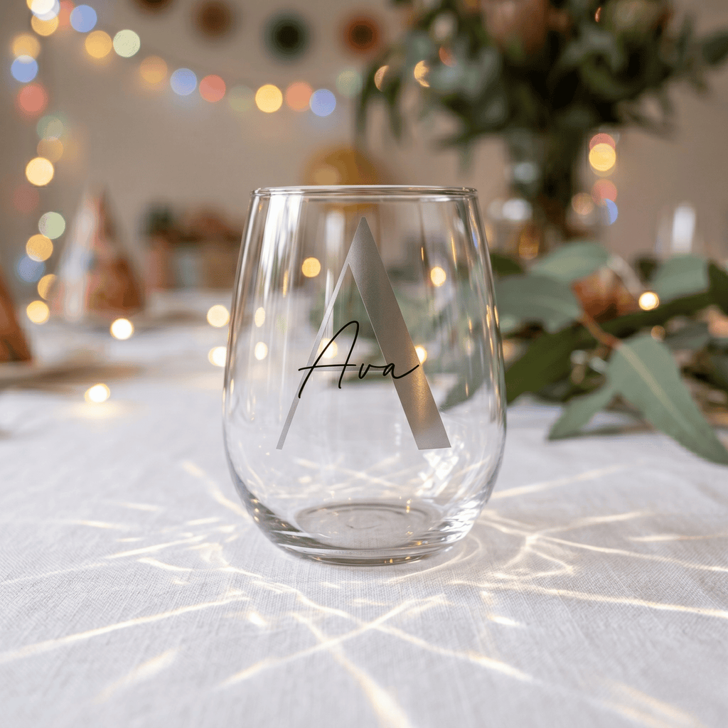 Clear glass with a monogram on a tablecloth with decorative lights and plants in the background
