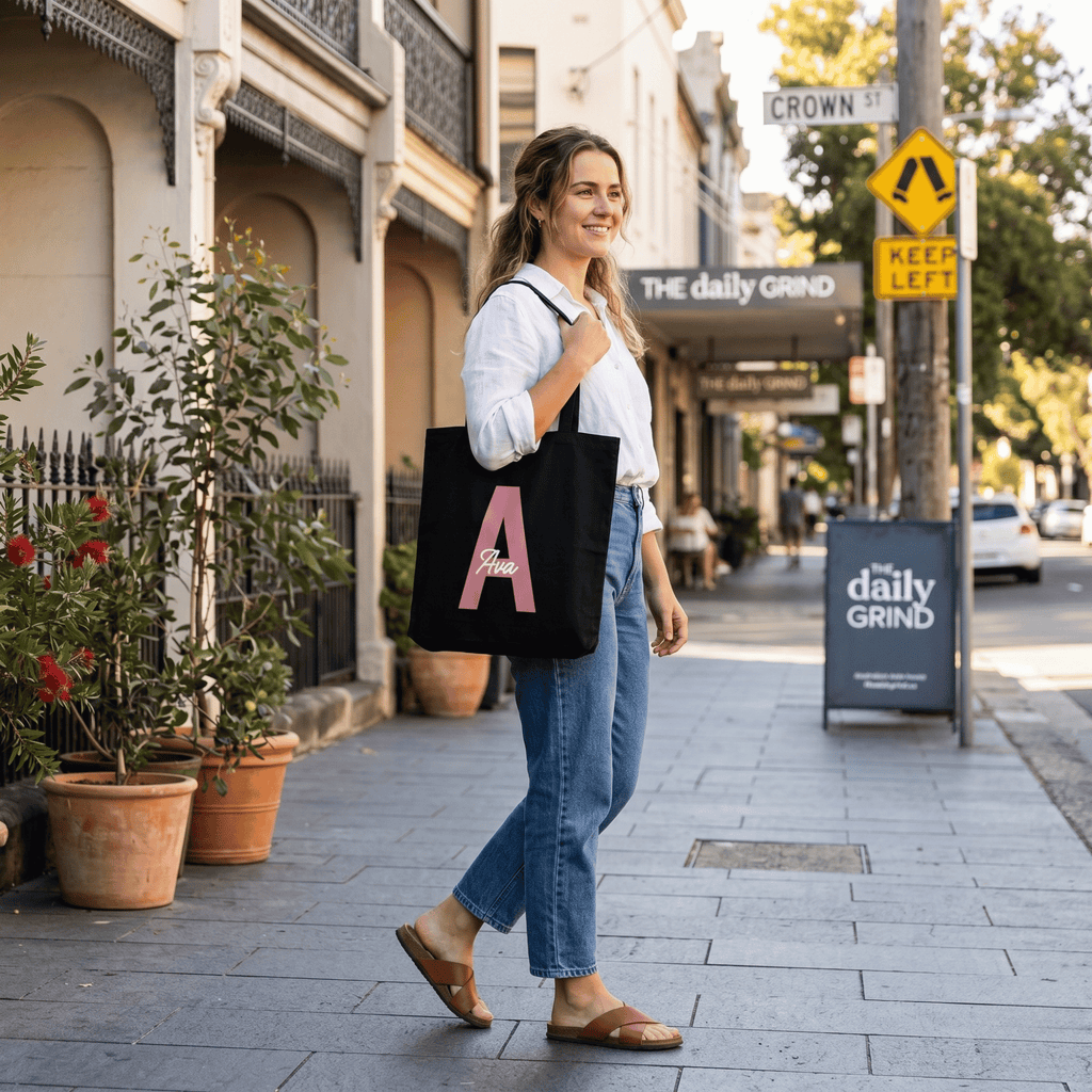 Woman walking on a street holding a black tote bag with a pink letter 'A' in an urban setting.
