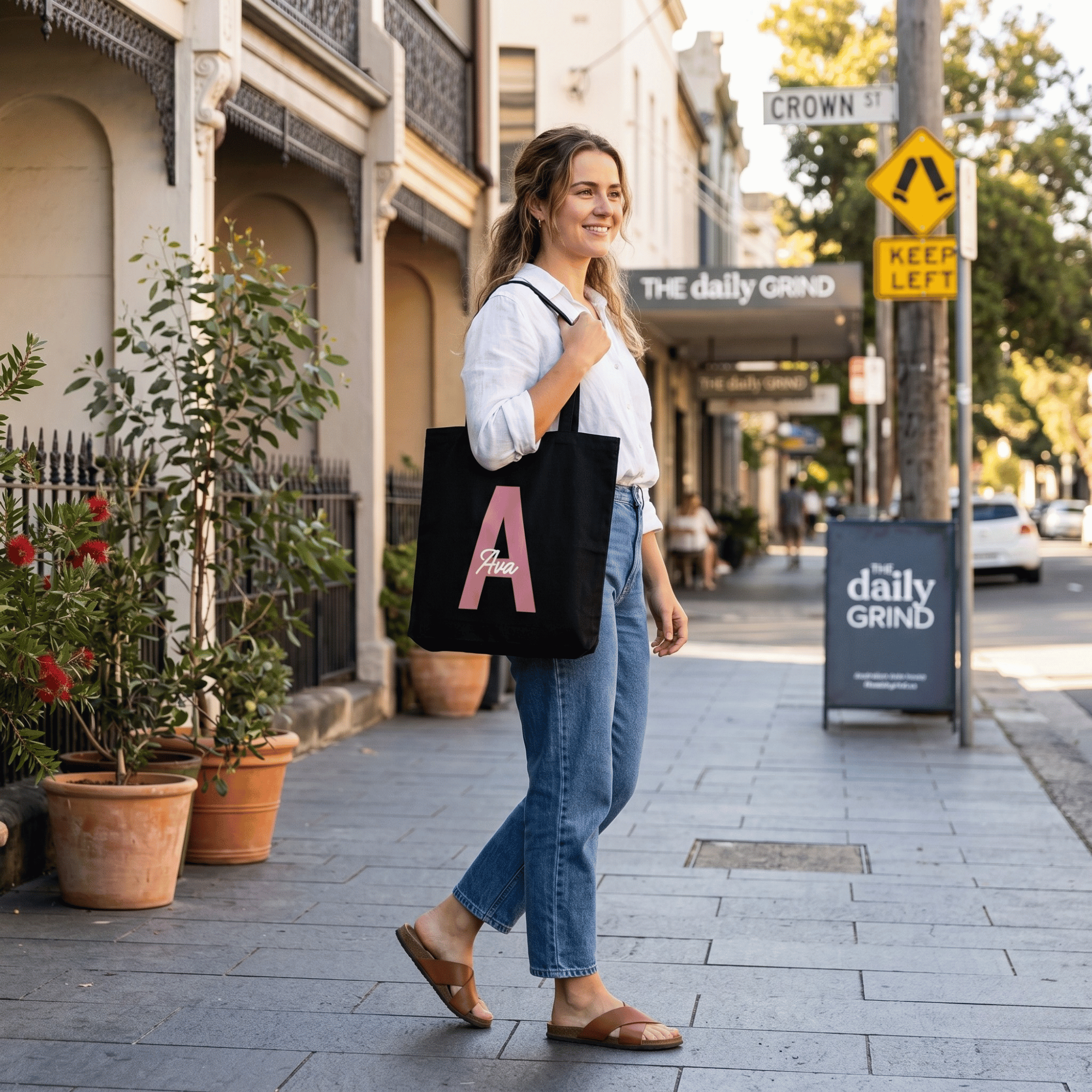Woman walking on a street holding a black tote bag with a pink letter 'A' in an urban setting.