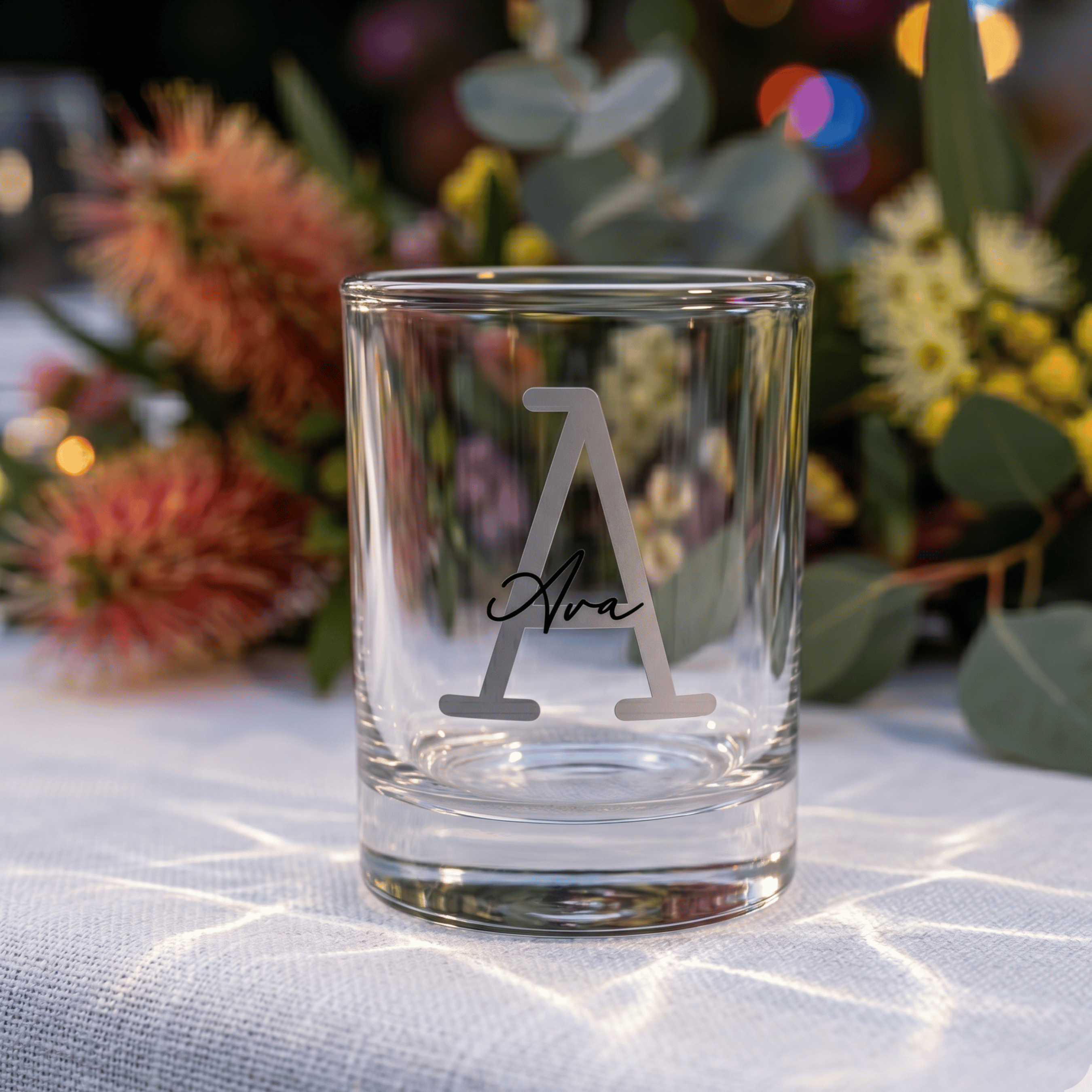 Clear glass with engraved design on a tablecloth with flowers in the background