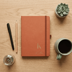 Orange notebook with 'Katie' on a wooden surface with a pen, coffee, and plant.