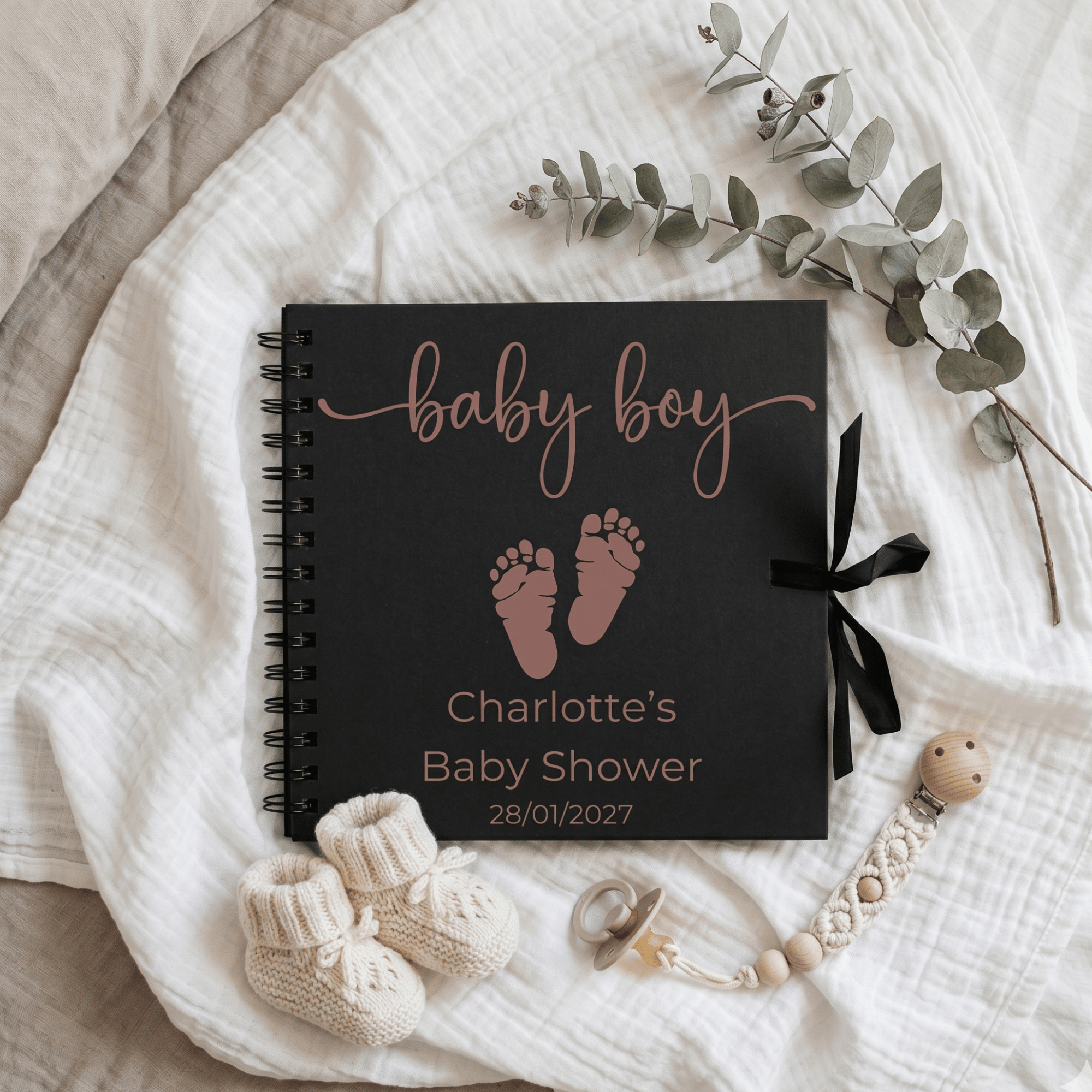 Black baby book with 'baby boy' text and baby shower details on a white blanket with decorative items.