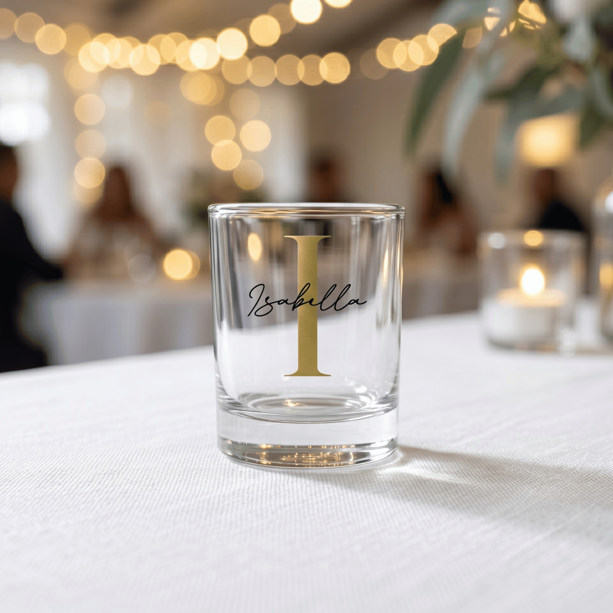 Clear glass with gold letter 'I' and name 'Isabella' on a white tablecloth with blurred lights in the background