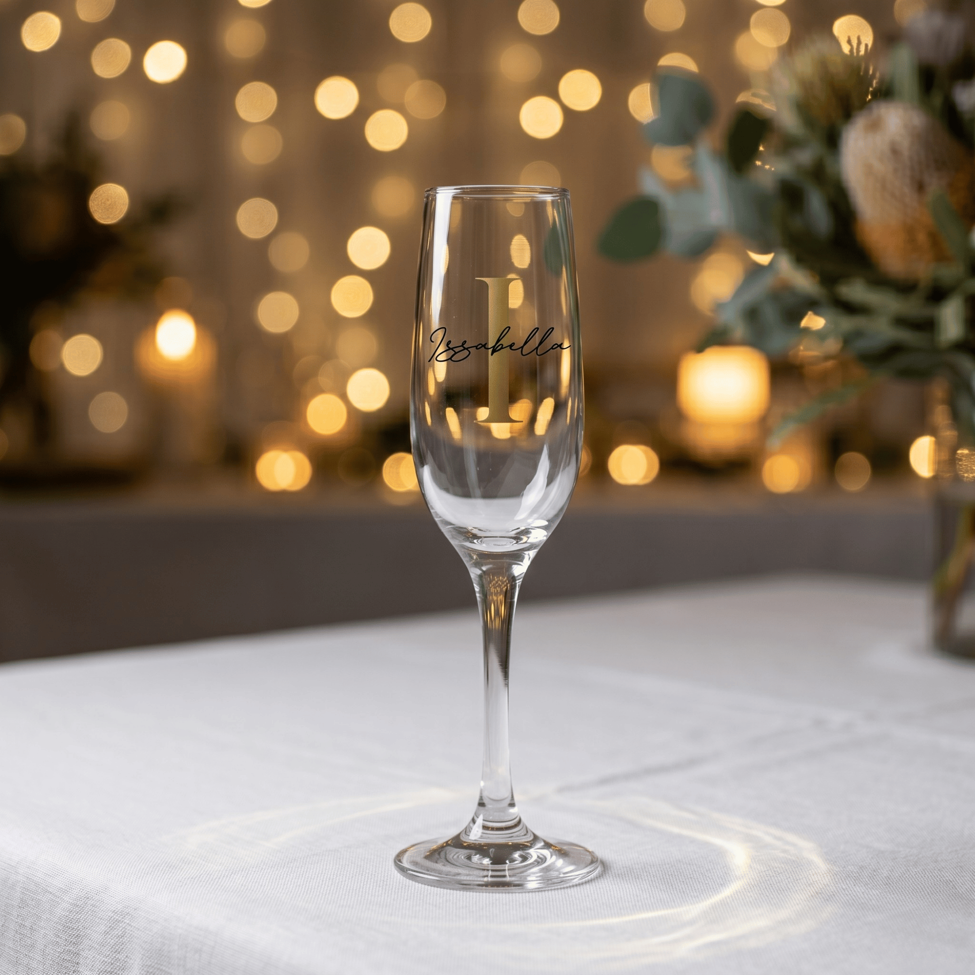 Champagne flute with 'L' monogram on a white surface with blurred lights and flowers in the background