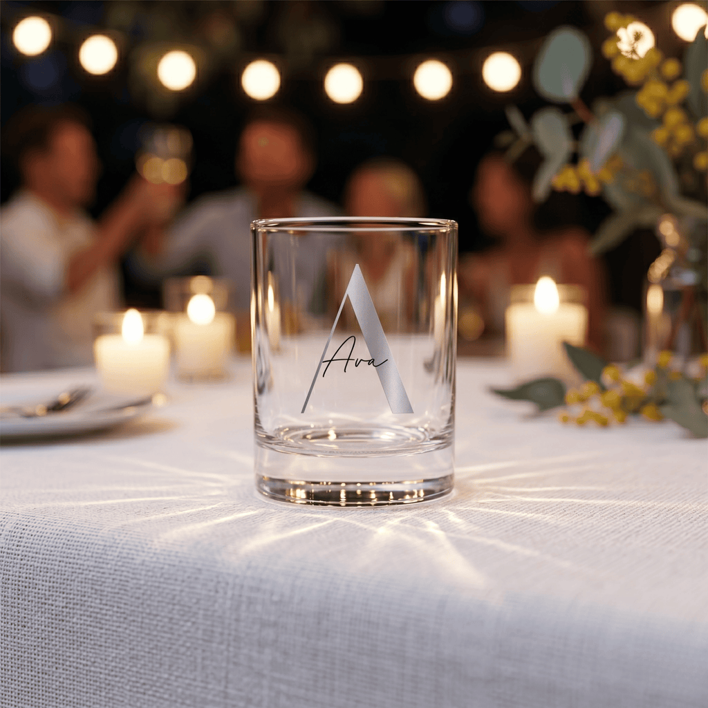 Clear glass with a monogram on a table with candles and people in the background