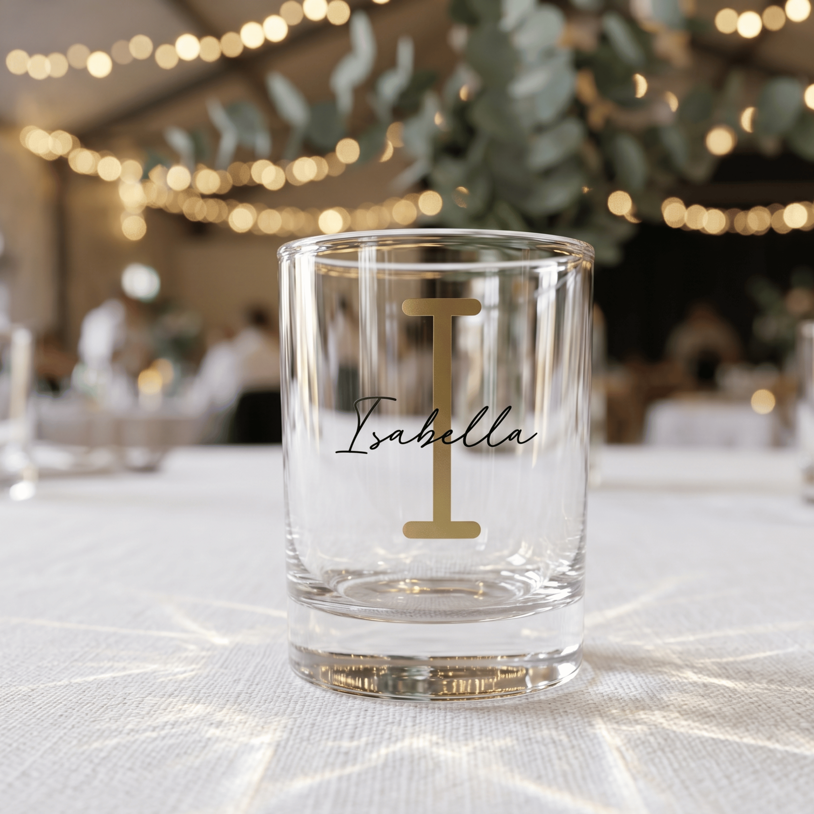 Clear glass with 'Isabella' and a gold monogram on a tablecloth with blurred festive background