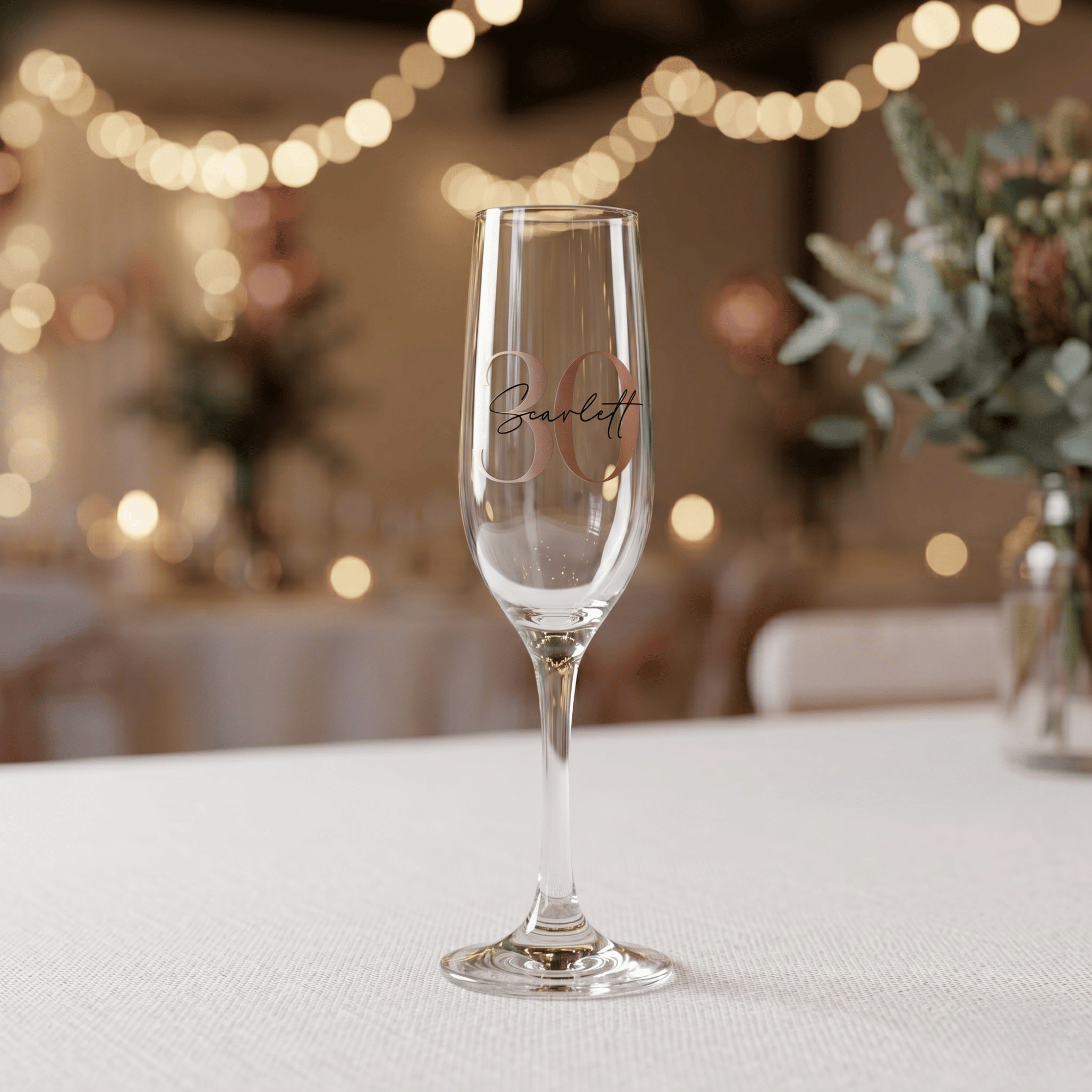 Champagne flute with 'Scarlett' engraved on a table with decorative lights and plants in the background
