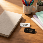Two name tags on a wooden surface with a notebook, mug, and plant in the background.