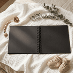 Black Album on a textured white surface with baby booties and a branch of leaves.