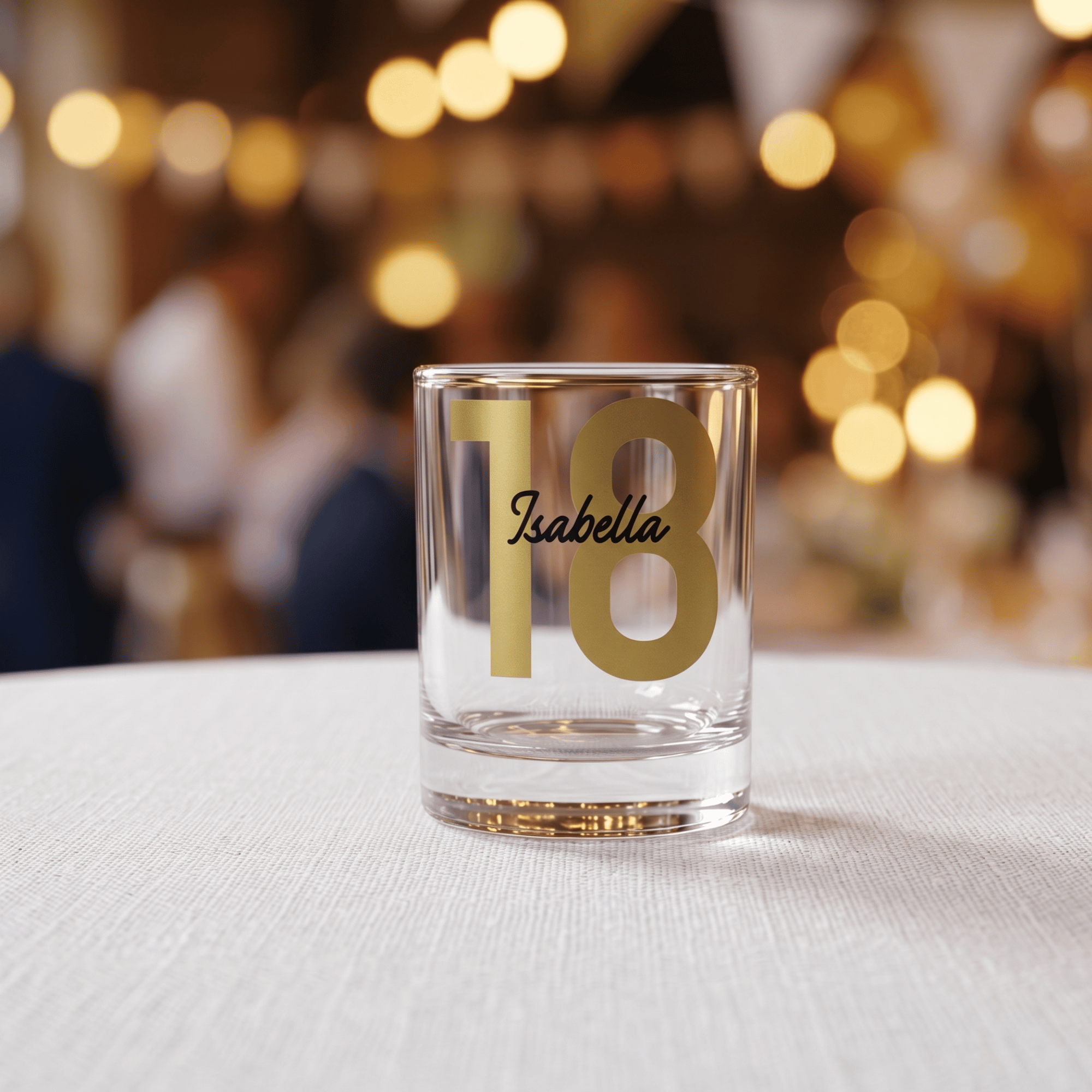 Clear glass with gold number '18' and name 'Isabella' on a blurred background