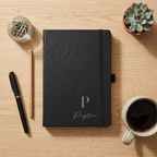 Black notebook with 'Peyton' on a wooden desk with a pen, coffee, and plant.