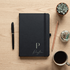 Black notebook with a monogram on a wooden table with stationery items.