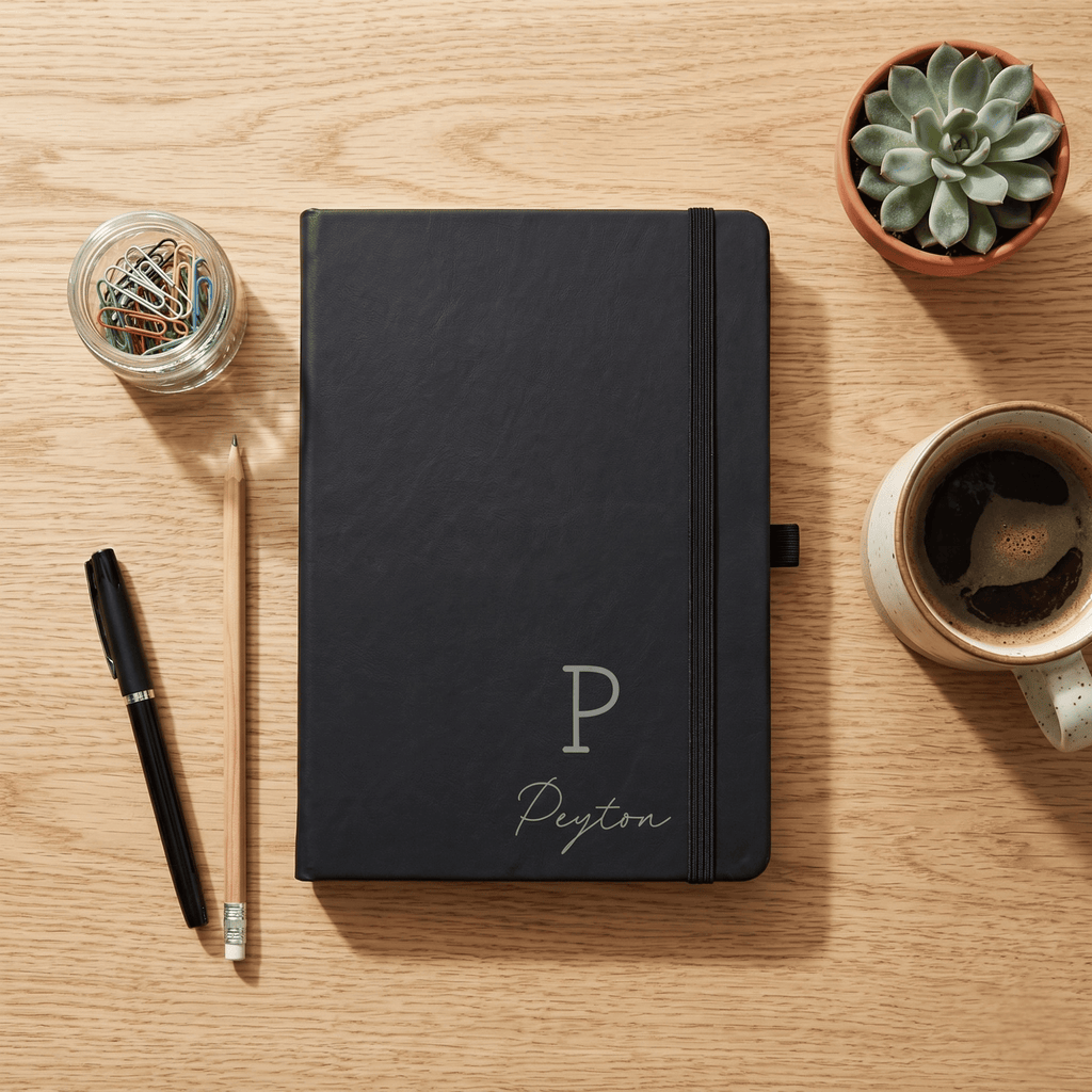 Black notebook with 'Peyton' on a wooden surface with a pen, coffee, and plant.