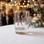 Clear glass with '18 Scarlett' printed on it, placed on a table with blurred background of people and lights.