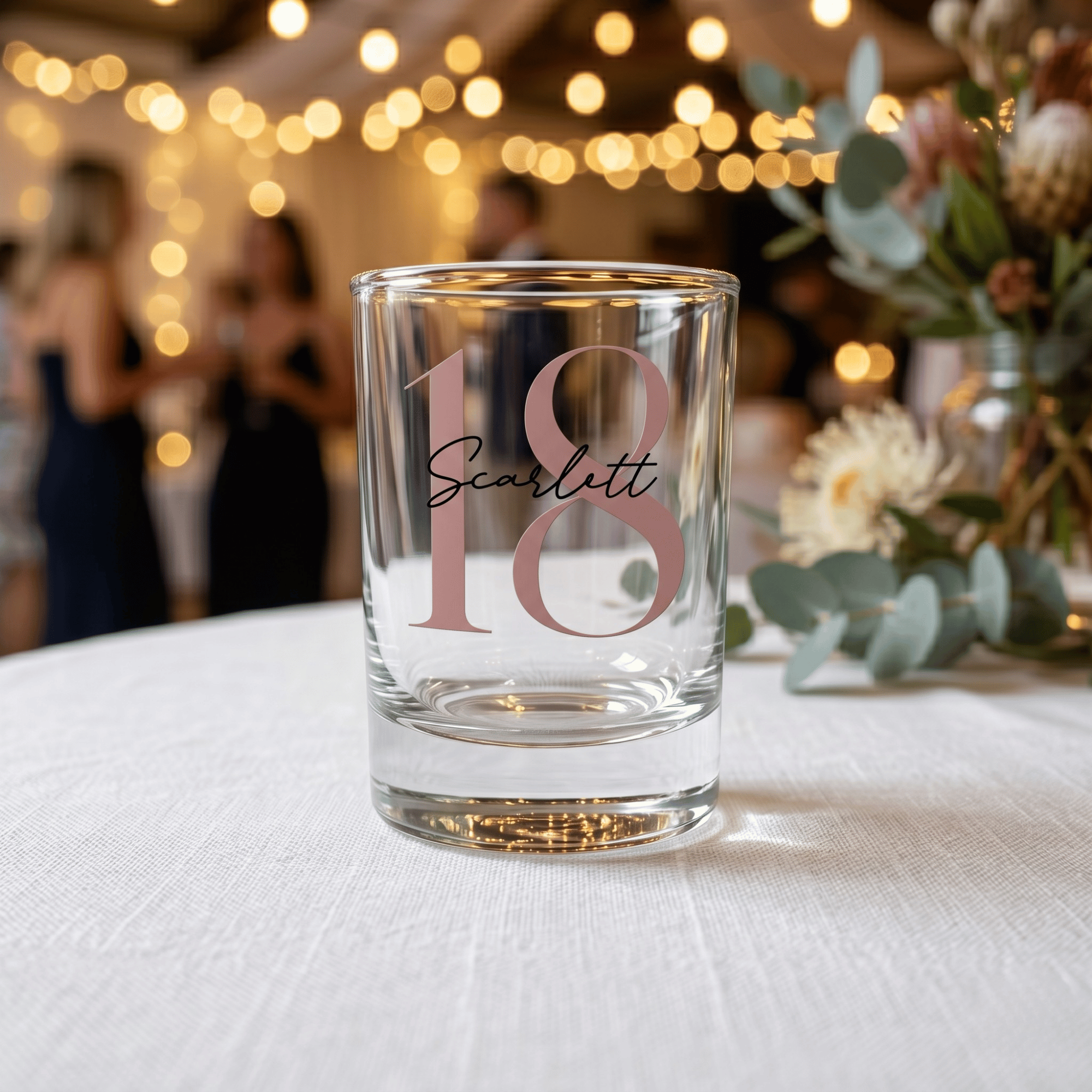 Clear glass with '18 Scarlett' printed on it, placed on a table with blurred background of people and lights.