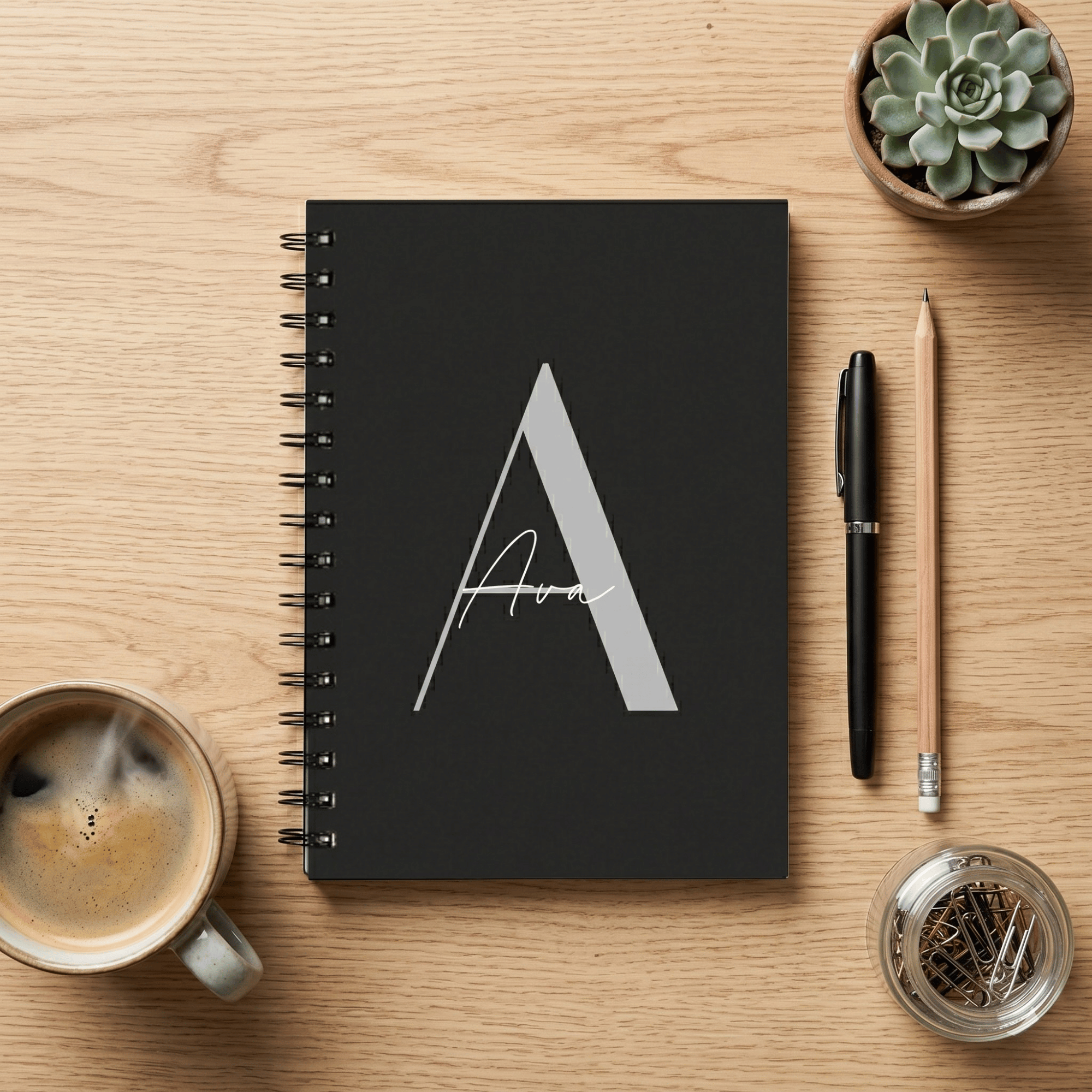 Black notebook with a white letter 'A' on a wooden table with a coffee cup, pen, and small plant.
