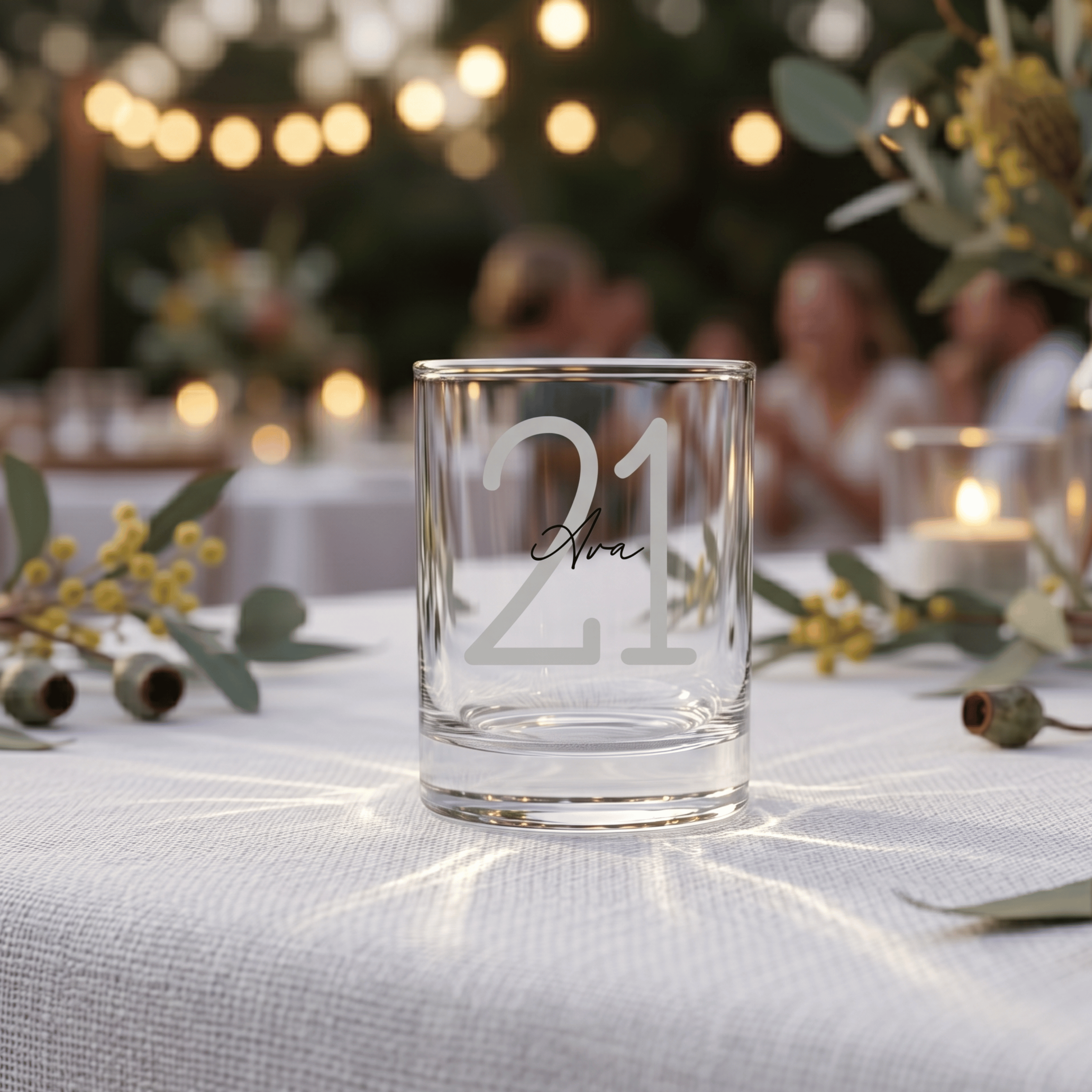 Clear glass with '21' engraving on a table with decorative elements and blurred people in the background