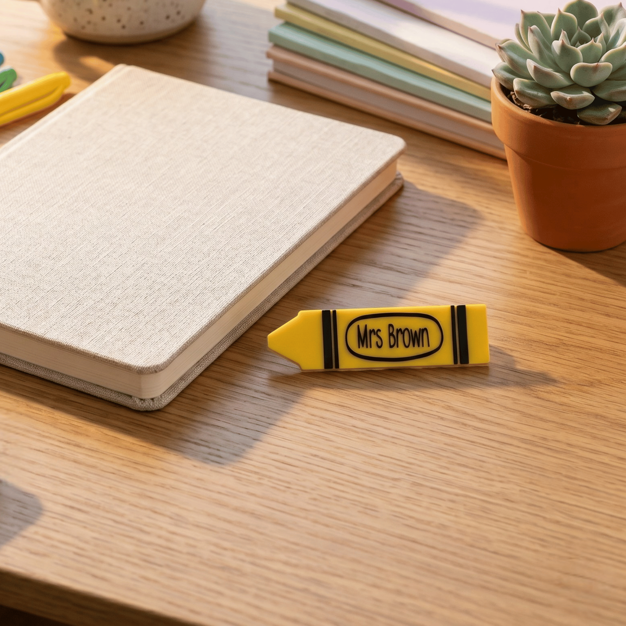 Yellow crayon with 'Mrs Brown' text on a wooden surface next to a notebook and plant.