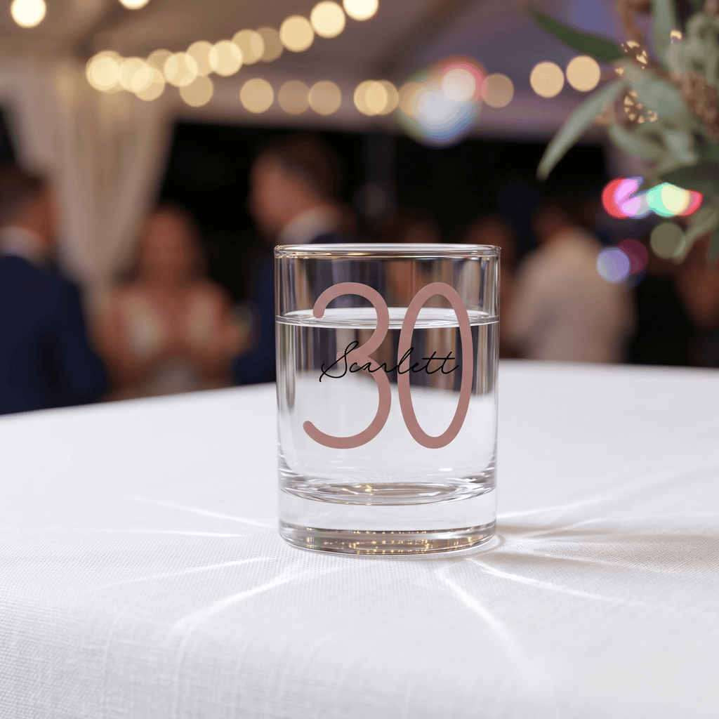 Clear glass with '30 Scarlett' on a white tablecloth with blurred background