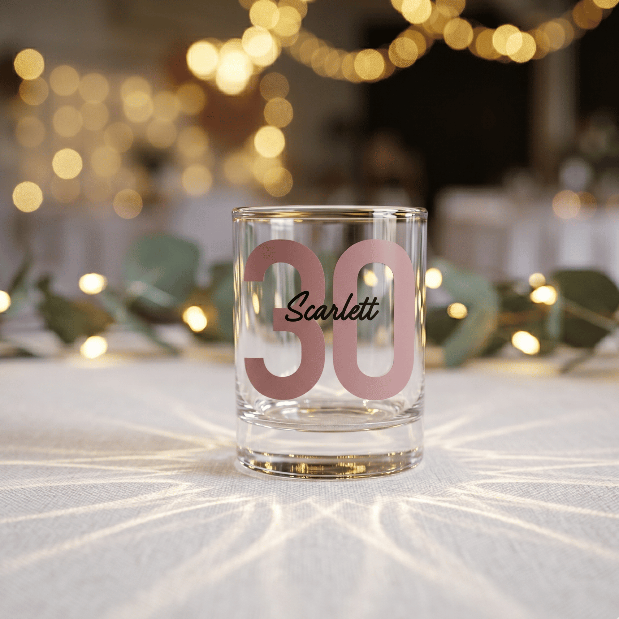 Clear glass with pink number '30' and name 'Scarlett' on a blurred festive background
